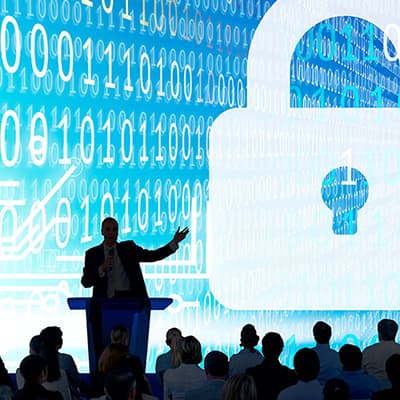 Speaker presenting cybersecurity awareness training to an audience with binary code backdrop.